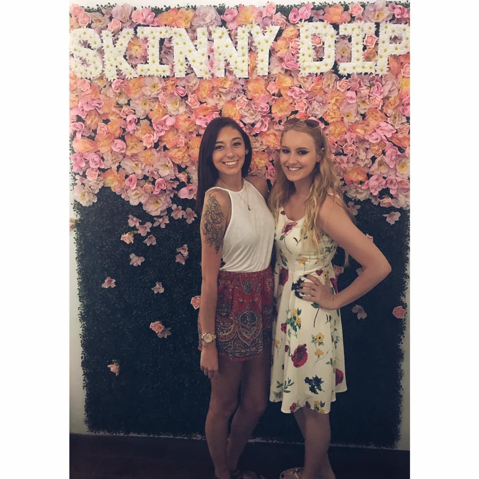a blonde and a brunette dressed in spring clothes standing together against a floral backsplash that spells out "skinny dip" the name of the coffee shop they are in.