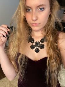 A selfie, Im a white blonde woman with long blonde hair that hits just at my waist. My hair is slightly crimped and my arm extends pulling a piece of hair away from my face. I have a soft white glow of eyeshadow around my blue eyes. I’m wearing a statement black vintage necklace and an a-line deep purple velvet dress.