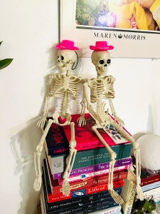 Two mini skeletons sit atop a colorful pile of books in matching pink Barbie cowboy hats